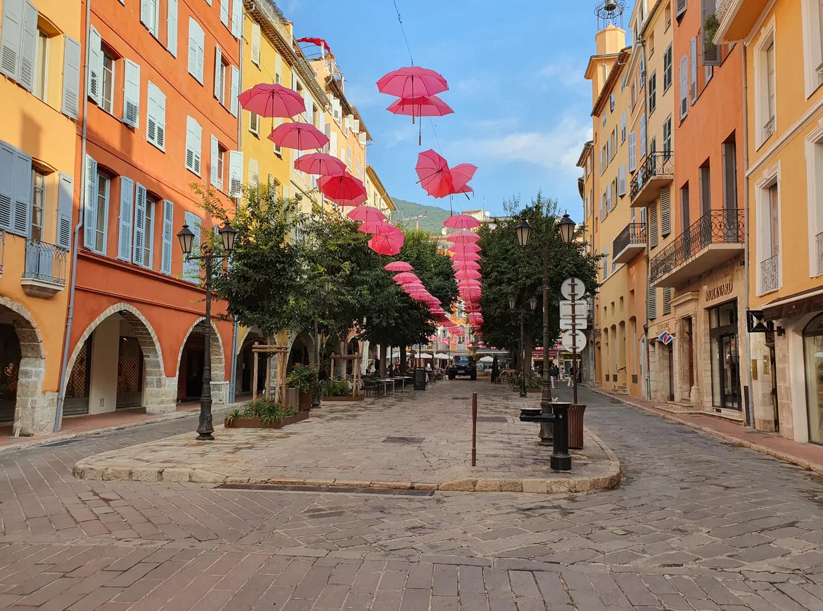 A cobbled lane in old Grasse lined with orange and yellow arcaded houses, with pink umbrellas hanging across the sky.