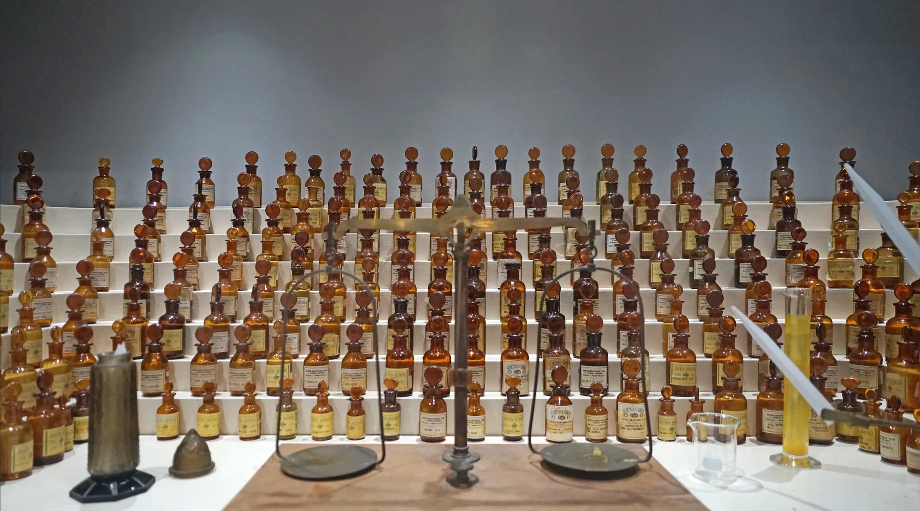 A tiered perfumer’s organ of amber glass bottles behind a brass balance and a measuring cylinder, in a museum in Grasse.