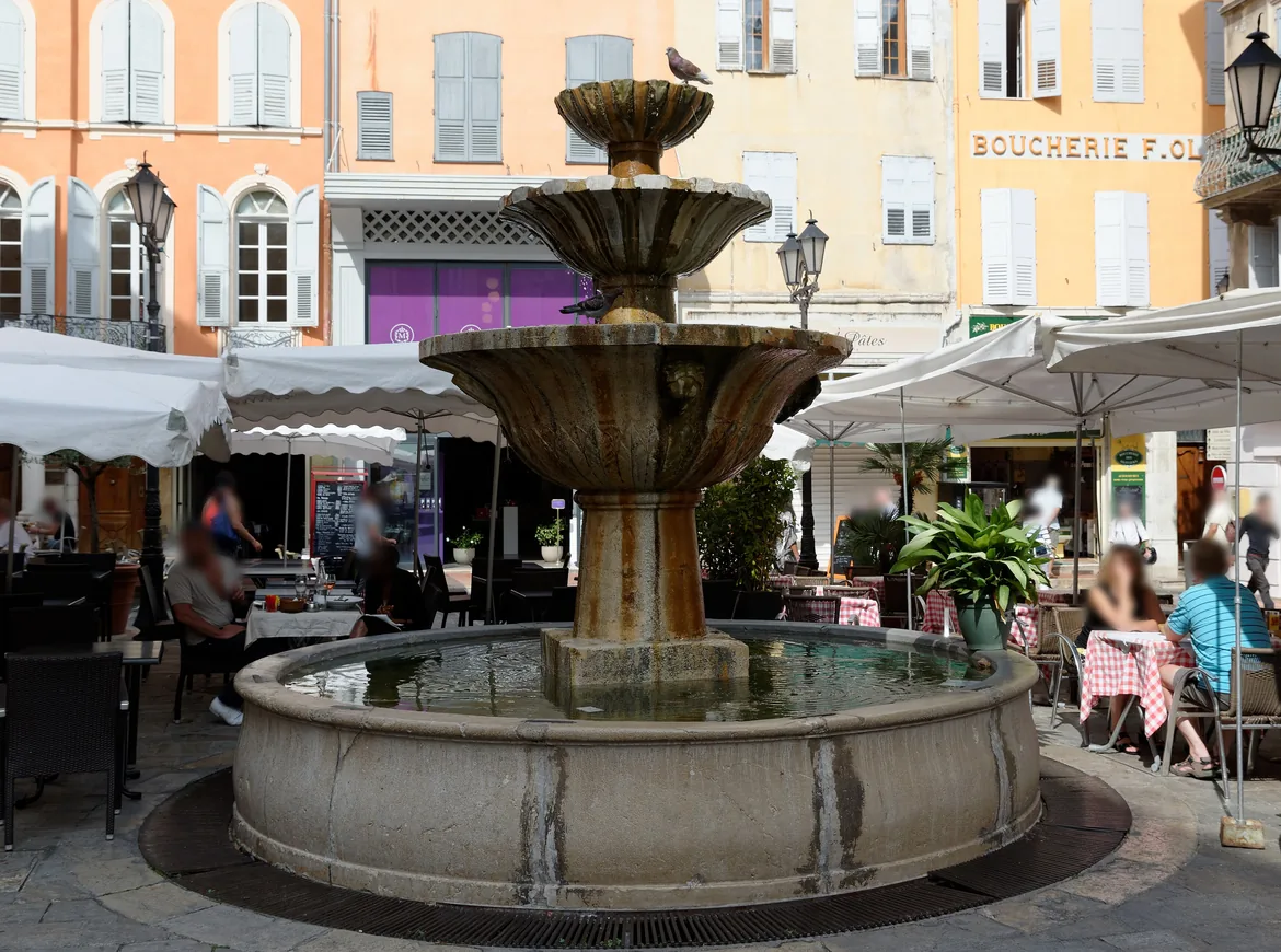 A three-tier stone fountain in a Grasse square, with café tables under white parasols and peach-coloured townhouses behind.